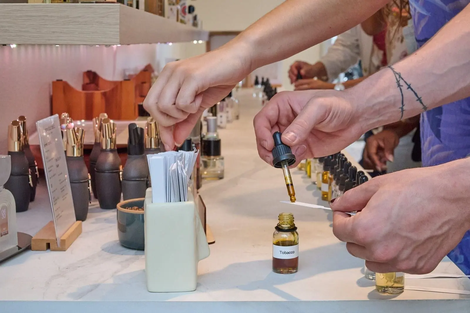 Hands dosing tobacco absolute onto a scent strip at the workshop table, beside a row of aroma materials